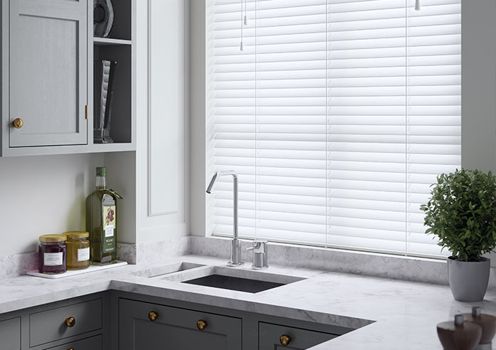 A white faux wooden blind adorns a window in a kitchen. A small plant is on the right of a marbled, grey-toned sink area.