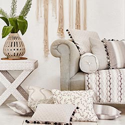 Cosy soft neutral cushions, on the floor next to a grey sofa. A cream vase with green plants sit on a table next to it.