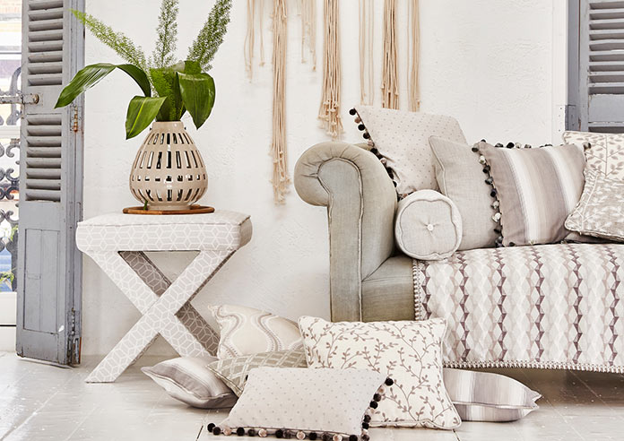 Cosy soft neutral cushions, on the floor next to a grey sofa. A cream vase with green plants sit on a table next to it.