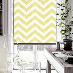A zigzag, striped yellow roller blind is shown in an modern home office. The desk is grey with a plant on it, and a clear seat is in the bnottom left hand corner.