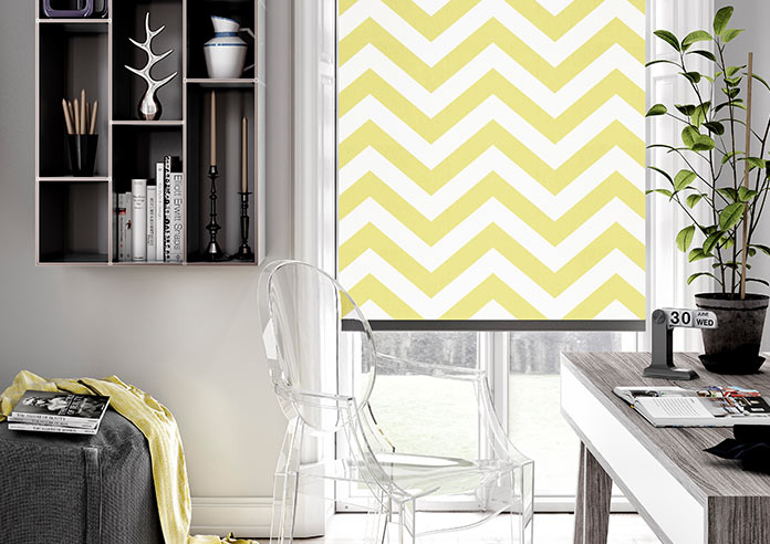 A zigzag, striped yellow roller blind is shown in an modern home office. The desk is grey with a plant on it, and a clear seat is in the bnottom left hand corner.