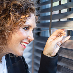 An image of amy wilson looking through her grey taped wooden blinds
