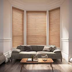 Real wooden blinds are shown in a living room bay window. The walls are cream, the sofa is grey and the room sparsely decorated.